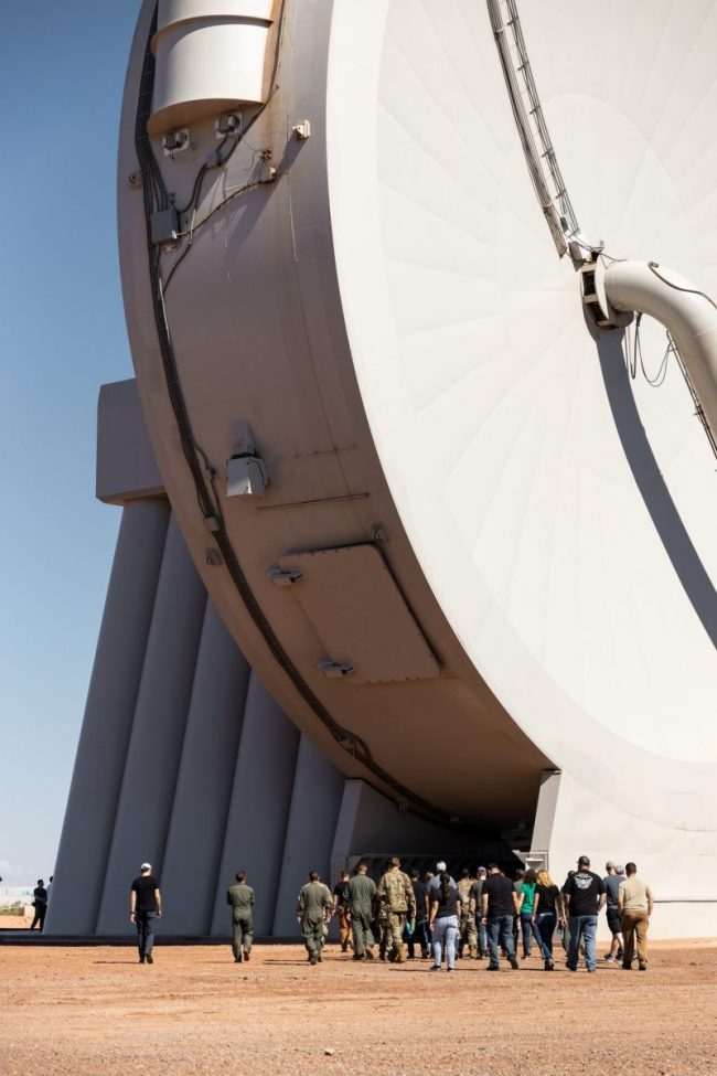 Launches: Group of people under large vertical disk shaped mechanical building.